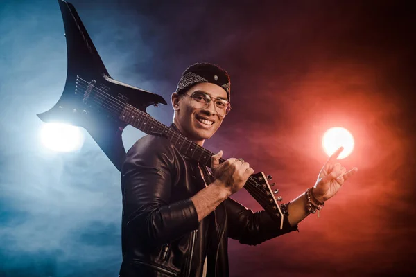 Rocker masculin en veste en cuir posant avec guitare électrique et faire des cornes signe sur scène avec de la fumée et un éclairage dramatique — Photo de stock