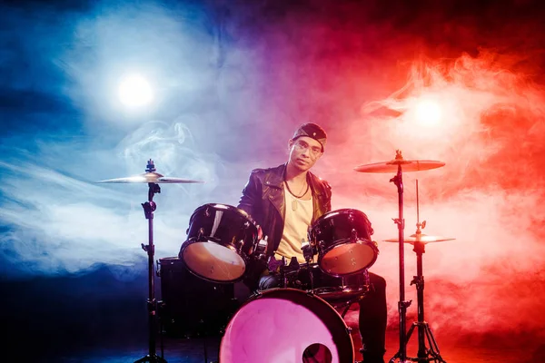 Confident mixed race male musician sitting behind drum set on stage with spotlights — Stock Photo