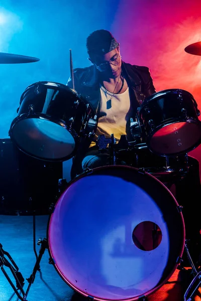 Handsome male musician in leather jacket playing drums during rock concert on stage with smoke and dramatic lighting — Stock Photo