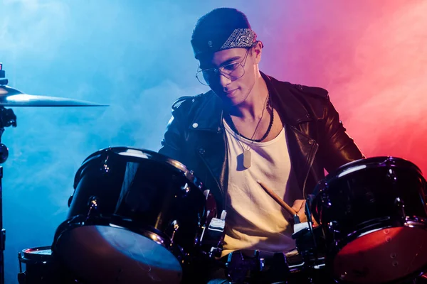 Joven de raza mixta tocando la batería durante el concierto de rock en el escenario con humo e iluminación dramática - foto de stock