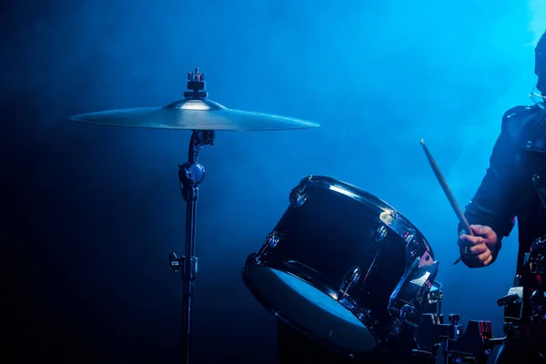 Частковий вид на чоловічого музиканта, який грає на барабанах під час рок-концерту на сцені з димом і драматичним освітленням — стокове фото