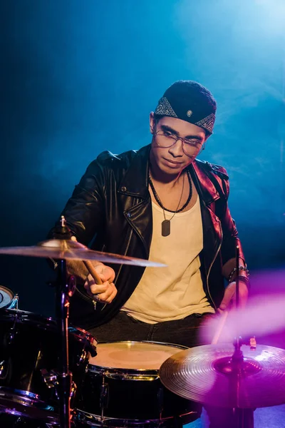 Joven músico joven y confiado en chaqueta de cuero tocando la batería durante el concierto de rock en el escenario con humo e iluminación dramática - foto de stock