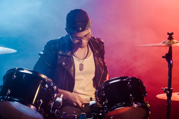 Sorridente jovem músico em jaqueta de couro tocando bateria durante concerto de rock no palco com fumaça e iluminação dramática — Fotografia de Stock