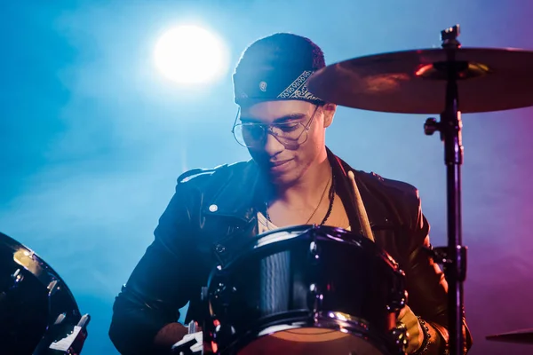 Uomo misto corsa in giacca di pelle suonare la batteria durante concerto rock sul palco con fumo e riflettori — Foto stock