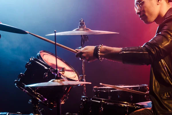 Musicista misto di razza maschile in giacca di pelle che suona la batteria durante il concerto rock sul palco con fumo e illuminazione drammatica — Foto stock