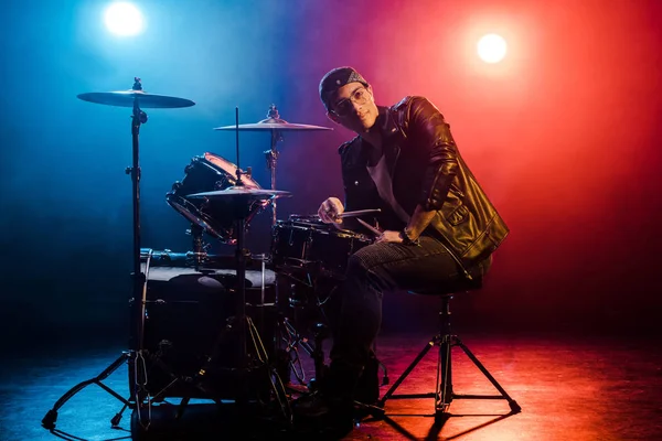 Young mixed race male musician sitting behind drum set on stage with spotlights and smoke — Stock Photo