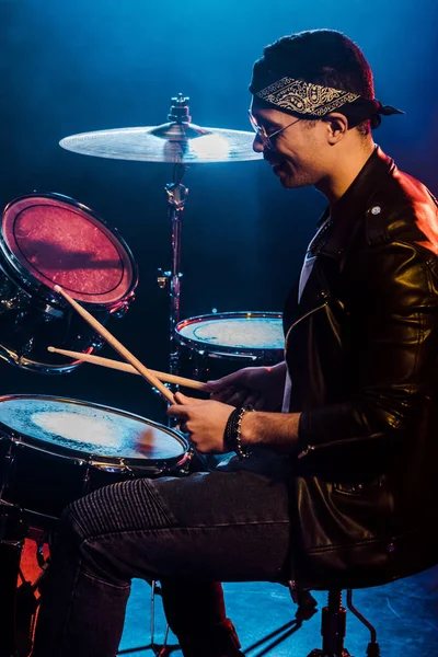 Happy male musician in leather jacket playing drums during rock concert on stage with smoke and dramatic lighting — Stock Photo