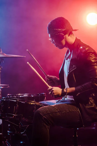 Вид збоку на чоловічого музиканта в шкіряній куртці, який грає на барабанах під час рок-концерту на сцені з димом і драматичним освітленням — стокове фото