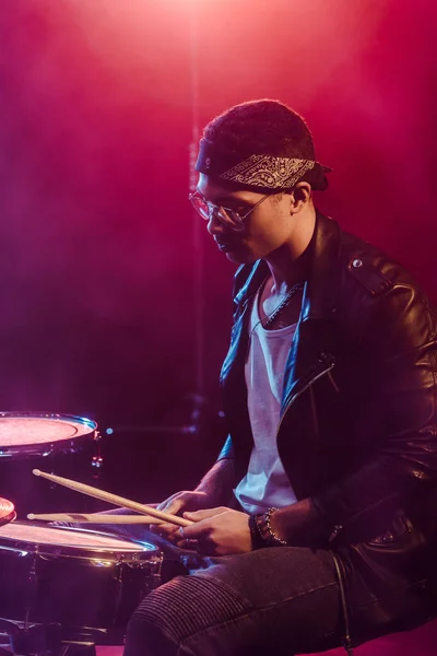Male musician in leather jacket playing drums during rock concert on stage with smoke and dramatic lighting — Stock Photo