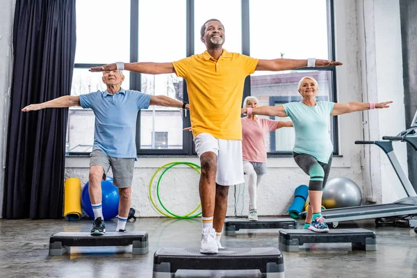 Atletas multiculturales mayores sonrientes ejercicio síncrono en plataformas de paso en el gimnasio - foto de stock