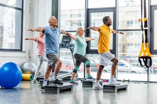 Esportistas seniores multiculturais sincrônicos exercitando-se em plataformas de degrau no ginásio — Fotografia de Stock