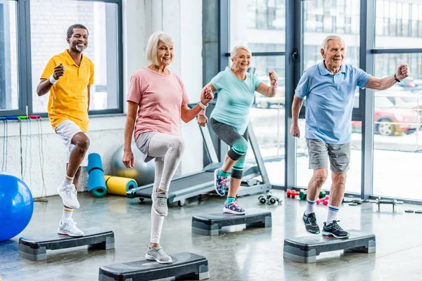 Atletas sénior multiétnicos sonrientes ejercicio síncrono en plataformas de paso en el gimnasio - foto de stock