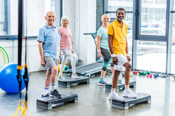 Atleti anziani multietnici esercizio sincrono su piattaforme passo in palestra — Foto stock