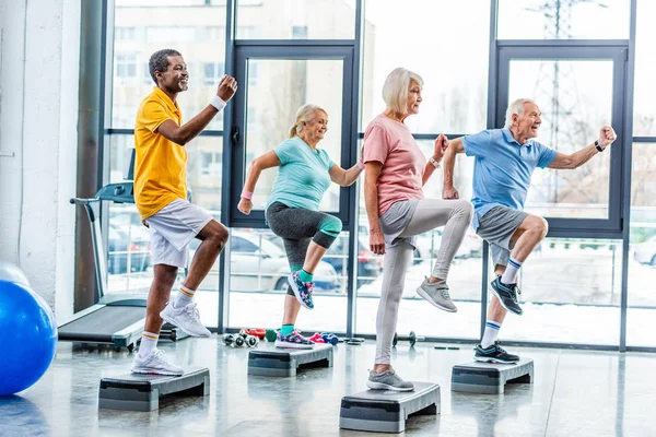 Vista laterale di atleti anziani multietnici esercizio sincrono su piattaforme passo in palestra — Foto stock
