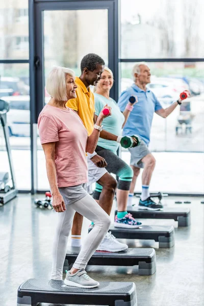 Side view of senior multicultural sportspeople synchronous exercising with dumbbells on step platforms at gym — Stock Photo