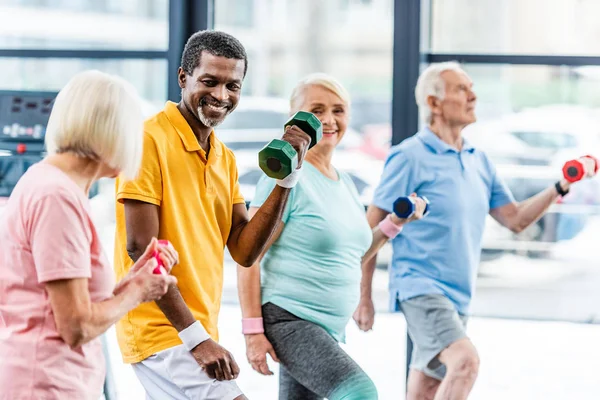 Hombre afroamericano riendo y sus amigos haciendo ejercicio con pesas en el gimnasio — Stock Photo