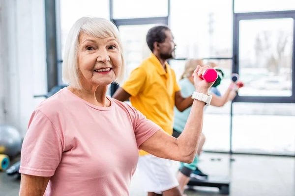 Selective focus of cheerful senior woman and her friends exercising with dumbbells at gym — Stock Photo