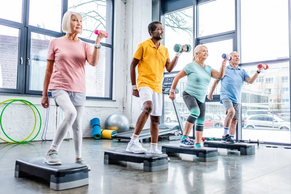 Desportistas seniores multiculturais sincrônicos exercitando com halteres em plataformas de degrau no ginásio — Fotografia de Stock