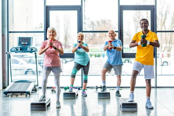 Sportifs multiculturels seniors exercice synchrone avec haltères sur les plates-formes étape à la salle de gym — Photo de stock