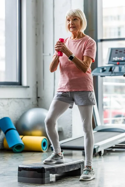 Deportista senior con smartwatch haciendo ejercicio con mancuerna en la plataforma de paso en el gimnasio - foto de stock