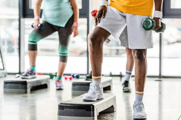 Imagen recortada del deportista afroamericano y sus amigos haciendo ejercicio con pesas en plataformas escalonadas en el gimnasio - foto de stock