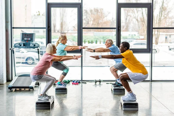 Atleti anziani multiculturali sincrono facendo squat su piattaforme passo in palestra — Foto stock