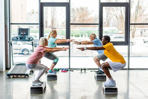 Atletas senior multiculturales sincrónicos haciendo sentadillas en plataformas de paso en el gimnasio - foto de stock