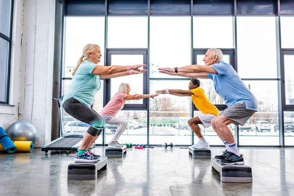 Vista lateral de los deportistas multiétnicos mayores felices sincrónicos haciendo sentadillas en plataformas escalonadas en el gimnasio - foto de stock