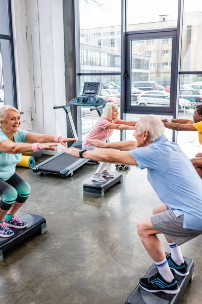 Allegro anziani sportivi multietnici sincrono facendo squat su piattaforme passo in palestra — Foto stock
