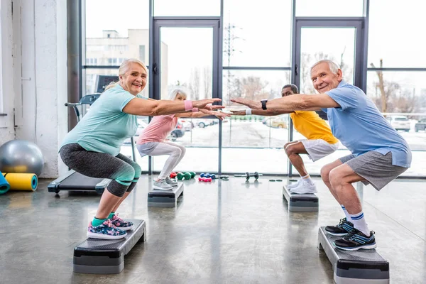 Glückliche Senioren multiethnische Sportler synchron machen Kniebeugen auf Trittbrettern in der Turnhalle — Stockfoto