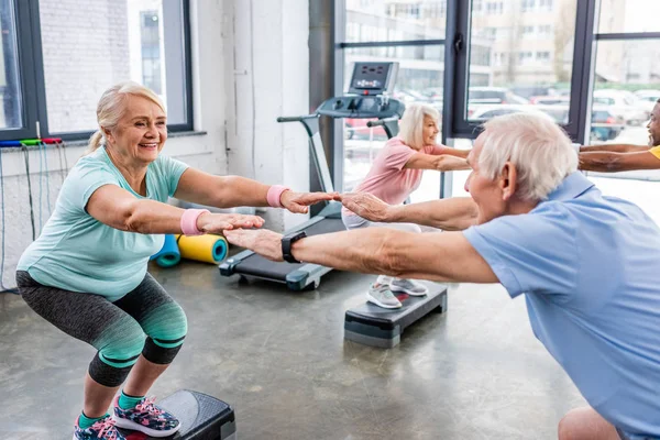 Selective focus of senior multiethnic sportspeople synchronous doing squats on step platforms at gym — Stock Photo