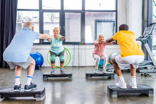 Seniors sportifs multiethniques synchrones faire squats sur les plates-formes pas à pas à la salle de gym — Photo de stock