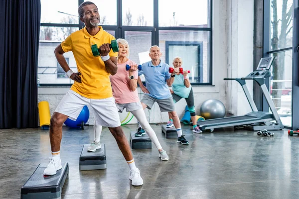 Multiculturale anziani sportivi sincrono esercizio con manubri su piattaforme passo in palestra — Foto stock