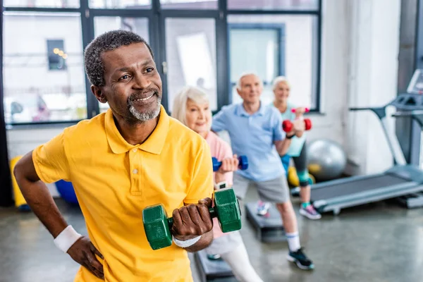 African american man and his friends synchronous exercising with dumbbells at sports hall — Stock Photo