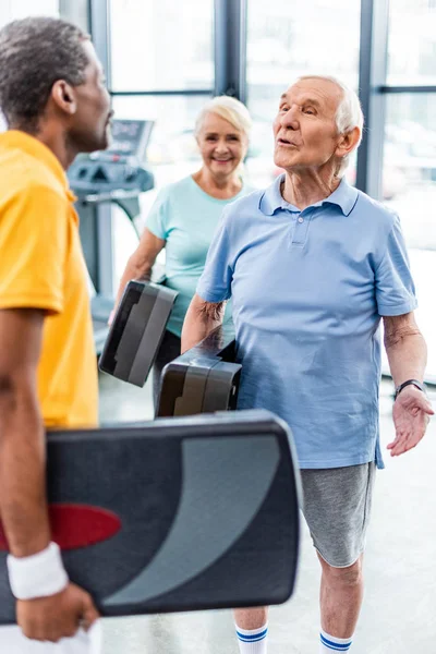 Two multiethnic male senior sportsmen with step platforms talking to each other at gym — Stock Photo