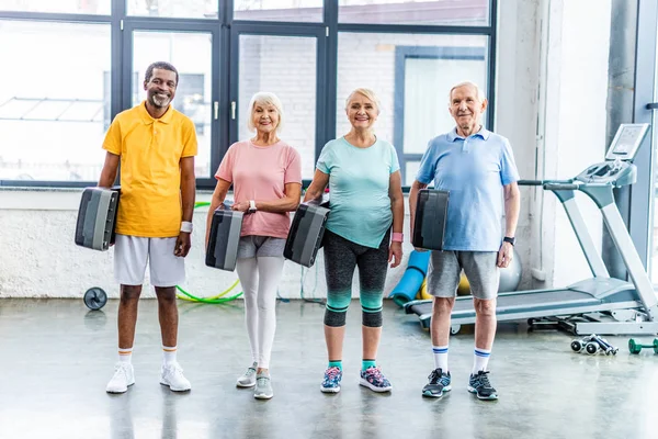 Cheerful multiethnic senior sportspeople holding step platforms at gym — Stock Photo