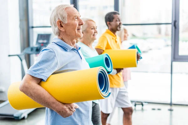 Side view of multethnic senior athletes holding fitness mats at sports hall — Stock Photo