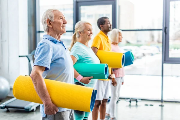 Vista laterale degli sportivi anziani multethnic che tengono tappeti fitness in palestra — Foto stock