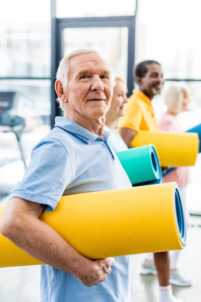 Selective focus of senior sportsman holding fitness mat and his friends standing behind at gym — Stock Photo