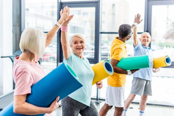 Sportifs multiethniques seniors souriants avec tapis de remise en forme prenant des hauts cinq les uns aux autres au gymnase — Photo de stock