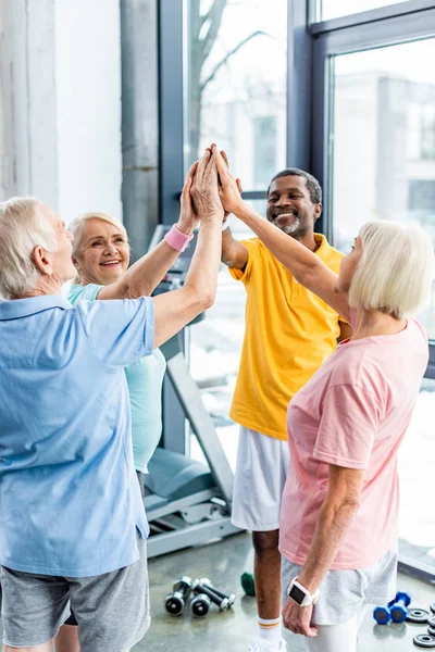 Sportifs multiculturels seniors souriants mettant la main ensemble à la salle de gym — Photo de stock