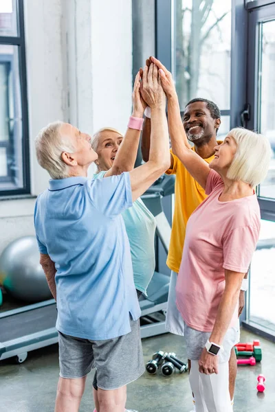 Sportifs multiculturels seniors souriants mettant la main ensemble à la salle de gym — Photo de stock