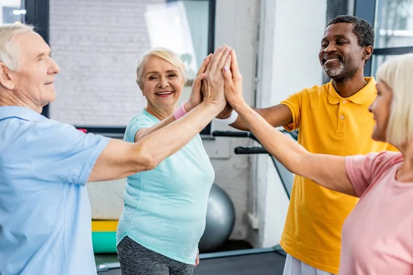 Souriant athlètes seniors multiculturels empilant les mains ensemble à la salle de gym — Photo de stock