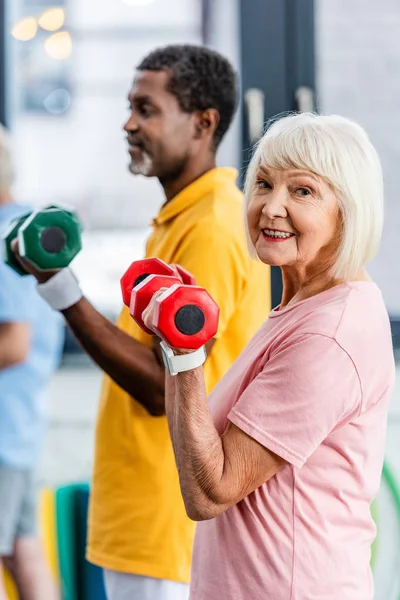 Selektiver Fokus der Seniorin und ihres afrikanisch-amerikanischen Ehemannes bei Übungen mit Hanteln im Fitnessstudio — Stockfoto