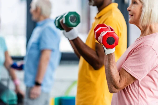 Vue partielle du couple mature interracial faisant de l'exercice avec haltères à la salle de gym — Photo de stock