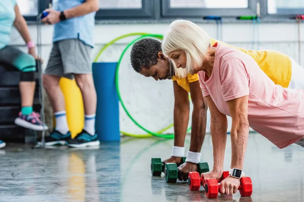 Selective focus of interracial couple doing plank with dumbbells at gym — Stock Photo