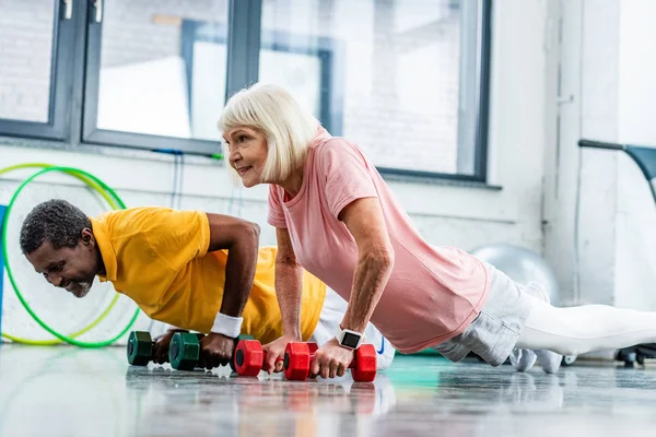 Pareja madura interracial haciendo flexiones con mancuernas en el gimnasio - foto de stock