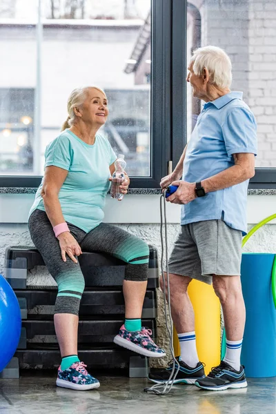 Sorridente coppia anziana con corda di salto e bottiglia d'acqua a riposo e parlando tra di loro in palestra — Foto stock