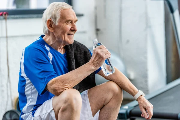 Sorrindo desportista sênior com toalha e garrafa de água descansando no ginásio — Fotografia de Stock