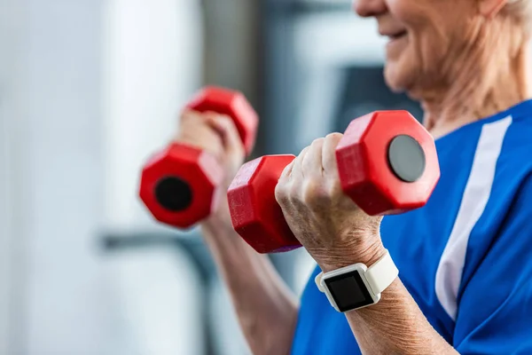 Visão parcial do desportista sênior com smartwatch fazendo exercício com halteres no ginásio — Fotografia de Stock